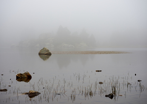 View of the middle of a lake on a foggy day, with a few rocks and vegatation standing in the middle, Lac des Bouillouses, Font-Romeu, France