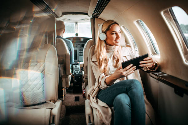 moda joven sentado en un avión privado y mirando por una ventana mientras escucha música a través de auriculares con una tableta digital - status symbol audio fotografías e imágenes de stock