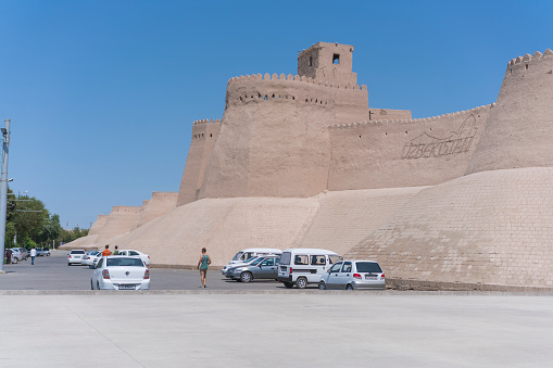 Khiva, Uzbekistan - August 05, 2018: Parking with cars and same tourists walking on it. In background is surrounding Wall of the old town of Khiva surrounding the Ancient city at the hat morning day in Uzbekistan.