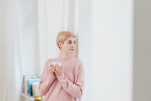 Pretty young blond woman relaxing with coffee standing leaning against a wall indoors looking back out of a curtained window