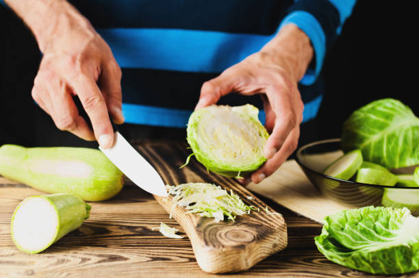 istock Man cuts fresh cabbage on cutting board on old brown wooden table with kitchenware on black background 1077164638