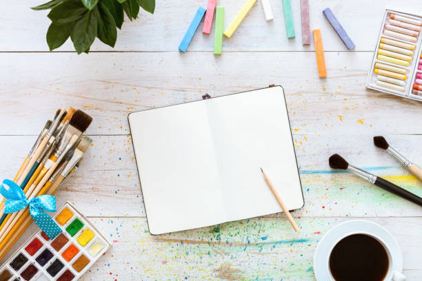 istock Colorful creative table with blank notebook for sketches and paints, pencil, paintbrushes set and cup of coffee on white wooden table, top view, copy space, flat lay style, drawing class education 1077171292