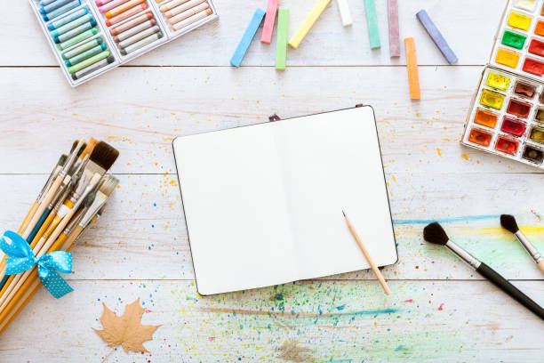 istock Artistic creative design workspace, notebook mock up for artwork with watercolor paints, pencil, set of paintbrushes on white wooden table, back to school, children kid art. Top view and copy space 1077171492