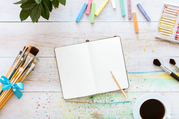 istock Creative art designer workplace, artistic mock up, blank notebook for sketches with pencil on it, set of paintbrushes and cup of coffee on white wooden table, top view, copy space, flat lay style 1077172006