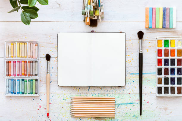 istock Creative work desk for self-expression, paint brushes, paintbox with watercolors, crayons, pencils and notebook paper on white wooden background, artistic workspace. Top view, flat lay, copy space 1077172390