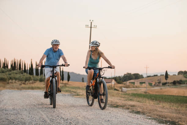 Senior Couple Cycling on Holiday Senior couple are cycling along a dirt road in Tuscany at sunset. man 60s bicycle stock pictures, royalty-free photos & images
