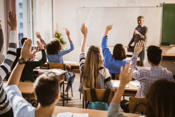 Back view of  high school students raising hands on a class. Rear view of large group of students raising their hands to answer the question on a class. high school building stock pictures, royalty-free photos & images