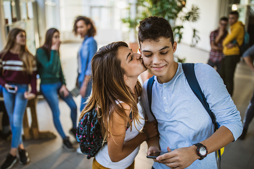 Loving female students kissing her boyfriend in the cheek at school hallway. There are people in the background.