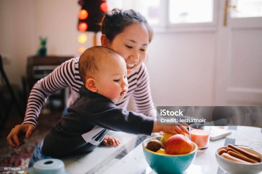 Young Asian mom giving food to her baby boy Brightly lit image of a young Thai mom with her little baby boy. They're spending time together at home, playing with toys and having some snacks. Asian family lifestyle concepts, shot in Europe. Eating Stock Photo