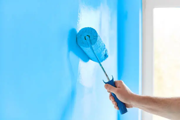 Photo of Male hand painting wall with paint roller. Painting apartment, renovating with blue color paint