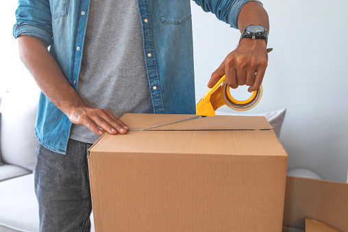 Close up of male hand packing cardboard box, concept moving house. House moving concept. Happy young couple moving into new apartment with packaging boxes. Male hands packing cardboard box with scotch
