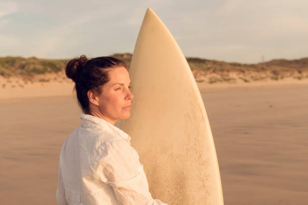 istock Woman standing holding a surfboard while looking at the view on Gwithian Towans Beach, Cornwall at near sunset. 1077275044