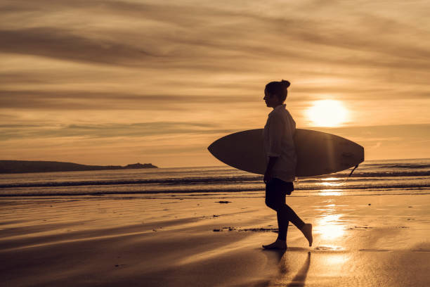 istock Backlit silhouetted woman with a surfboard at sunset on Gwithian Towans Beach, Cornwall on a June evening. 1077275064