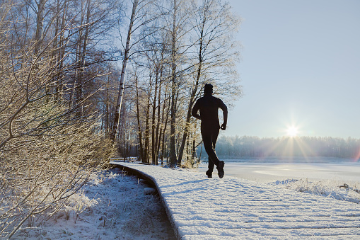 Young, adult alone man in tracksuit and hat running on forest trail in sunny winter morning after first snow. Enjoying sport in sunrise. Daily active lifestyle. Life motion. Back view.