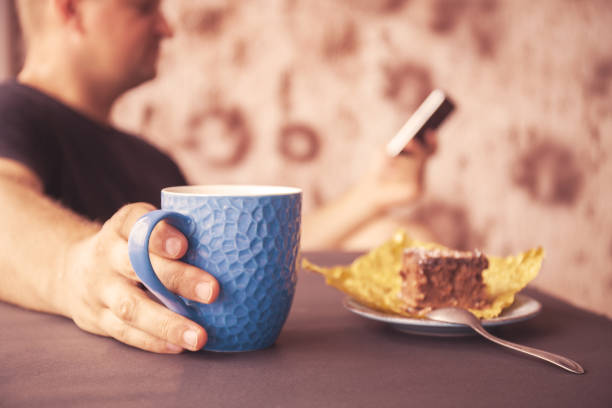 jovem, lendo uma revista em um café. em uma mão uma xícara de café, no outro um dispositivo eletrônico. olhando para um telefone móvel - men reading outdoors book - fotografias e filmes do acervo