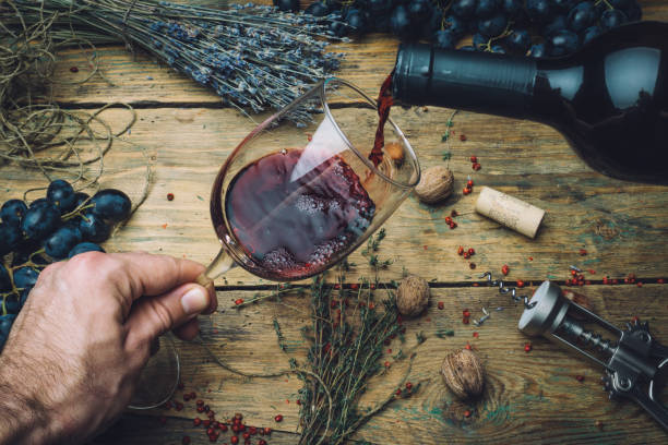 vigneron, verser le vin rouge (bio) pour la dégustation. rouge vin dégustation (vin bio) dans un verre de vin avec des raisins, noix et fines herbes sur le fond de l’ancienne table en bois. réglage de la table. - merlot cabernet photos et images de collection