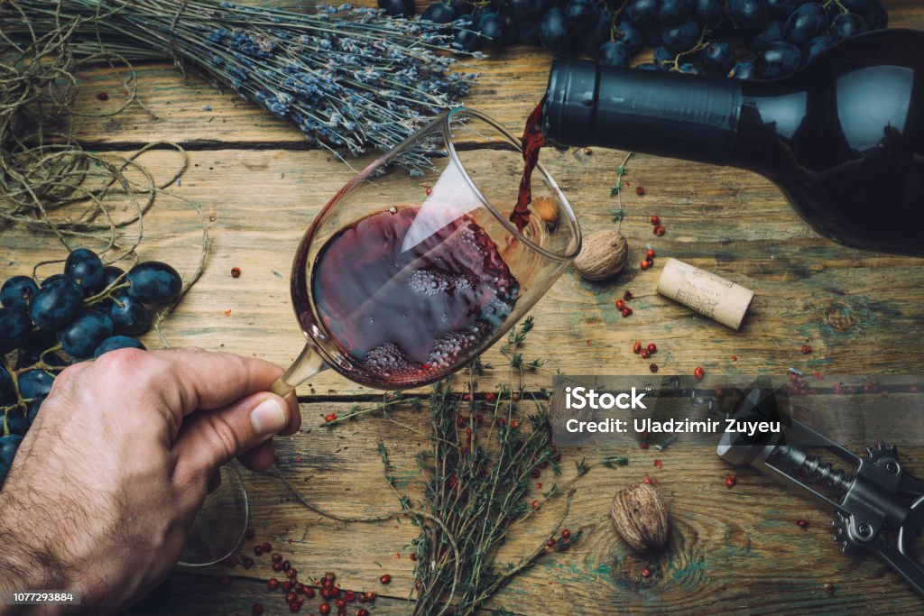 Wine maker pouring red wine (bio) for tasting. Red wine tasting (bio wine) in a wine glass with grapes, nuts and herbs on the background of the old wooden table. Table setting. Wine maker pouring red wine (bio) for tasting. Red wine tasting in a wine glass with grapes, nuts and herbs on the background of the old wooden table. Table setting. Wine Tasting Stock Photo