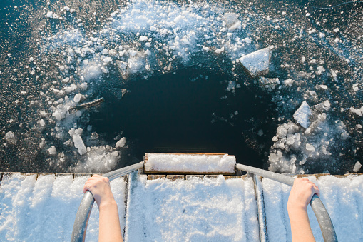 Young woman's hands holding on metallic ladder handles. Ready for swimming in ice hole at lake after sauna in cold day. Care about body health in winter time. Point of view shot. Close up.