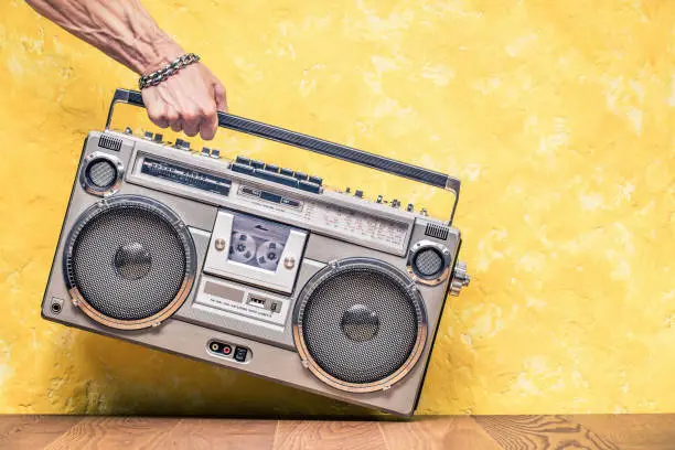 Retro outdated portable stereo boombox radio receiver with cassette recorder from circa 1980s in a strong man's hand front concrete textured yellow wall background. Vintage old style filtered photo