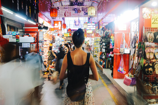 Young traveller woman walking through the small street markets in Singapore. She is carrying a backpack, strolling through the market stalls, looking for exotic and cheap stuff to buy, from technics and gadgets to delicious Asian street food.