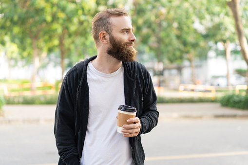 Pensive bearded man walking in city and holding plastic cup. Handsome guy standing, holding drink and looking away. Tourism and relaxation concept. Front view with trees in background.