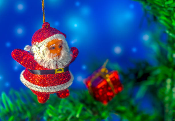 Santa Claus ornament on Christmas tree, close-up. Blurred white lights on blue color background. Blurred red color gift box on pine tree brunch.