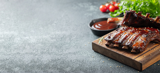 Closeup of pork ribs grilled with BBQ sauce and caramelized in honey. Tasty snack to beer on a wooden Board for filing on dark concrete background. With copy space.