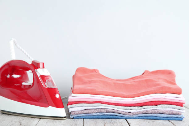 istock Stack of folded clothes with electric iron on gray background 1077558002