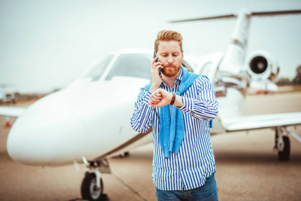 Young successful man getting out of a private jet parked on a runway. He is using a smart phone. He is also checking the time on his wristwatch.