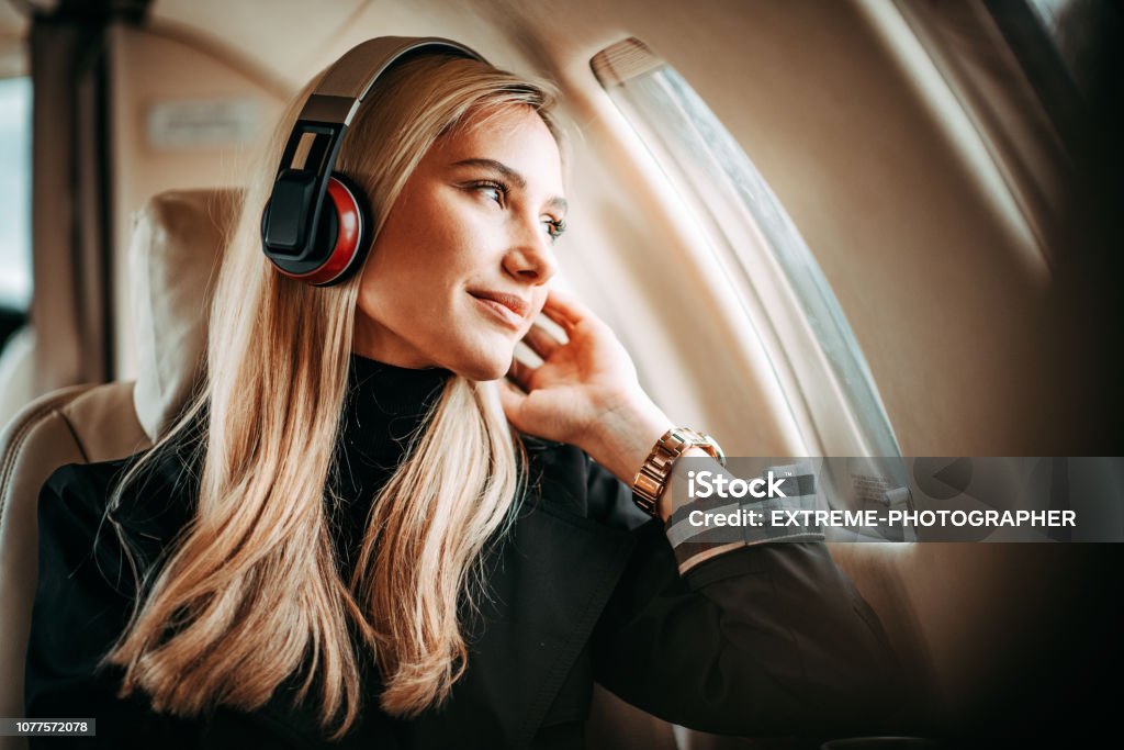 Beautiful young woman listening to music through the headphones in a private jet Young successful blonde woman sitting on a private jet and listening to music through headphones. Airplane Stock Photo