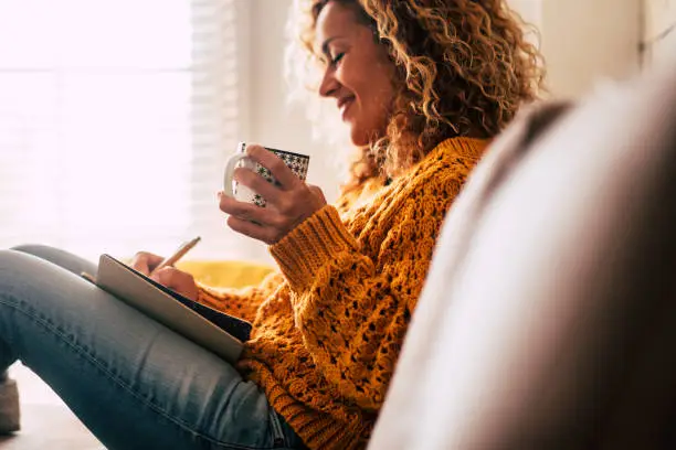 Photo of Happy cute lady at home write notes on a diary while drink a cup of tea and rest and relax taking a break. autumn colors and people enjoying home lifestyle writing messages or lists. Blonde curly beautiful lady sit down in the house