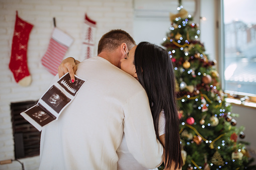 Man and woman, married couple relaxing together, hugging next to a Christmas tree, woman is holding ultrasound picture.