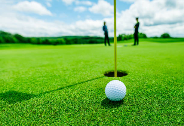golf ball in the green grass, almost in the hole and two blurred golf players golf ball in the green grass close to the hole with the shadow of the flagpole and two blurred guys in the background golf course photos stock pictures, royalty-free photos & images
