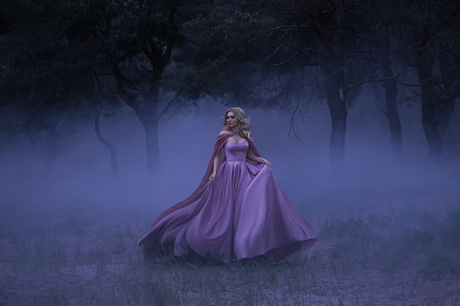 A frightened girl blonde runs away from a forest that has covered a thick fog. On the elf, a luxurious pink dress with a long train and a raincoat. Artistic photo