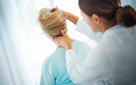 Closeup rear view of an early 60's senior lady having some neck pain due to dislocation and a light form of arthritis. Female doctor is examining her neck vertebrae by touching and slightly moving patient's head.