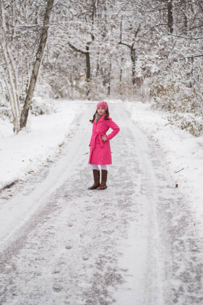12 year old girl with brown hair and brown eyes wearing pink coat and brown boots and fuzzy scarf and flower knit headband standing in the snow by trees.