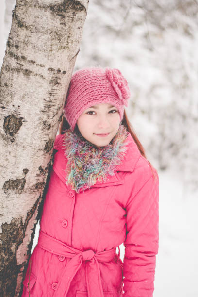12 year old girl with brown hair and brown eyes wearing pink coat and brown boots and fuzzy scarf and flower knit headband standing in the snow by trees.
