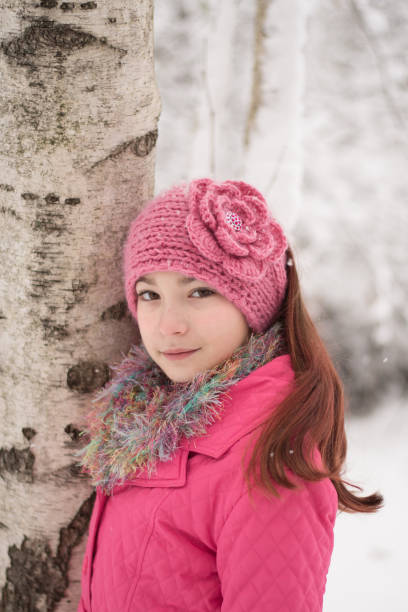 12 year old girl with brown hair and brown eyes wearing pink coat and brown boots and fuzzy scarf and flower knit headband standing in the snow by trees.
