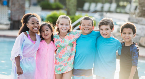 Six multi-ethnic children standing at swimming pool A group of six multi-ethnic children, 6 to 9 years old, standing side by side in front of a swimming pool, smiling at the camera. male swimsuit standing arm around stock pictures, royalty-free photos & images