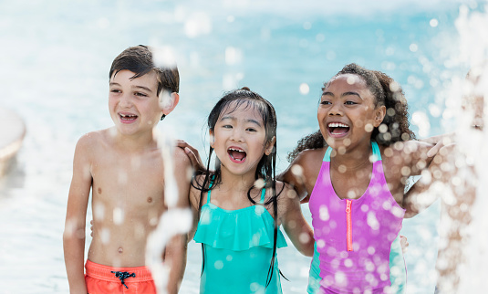 A group of three multi-ethnic children, 6 to 9 years old, having fun at a pool party. The Asian girl in the middle is laughing and shouting, looking toward the camera, standing between her two friends. A swimming pool is in the background and water is splashing around them.