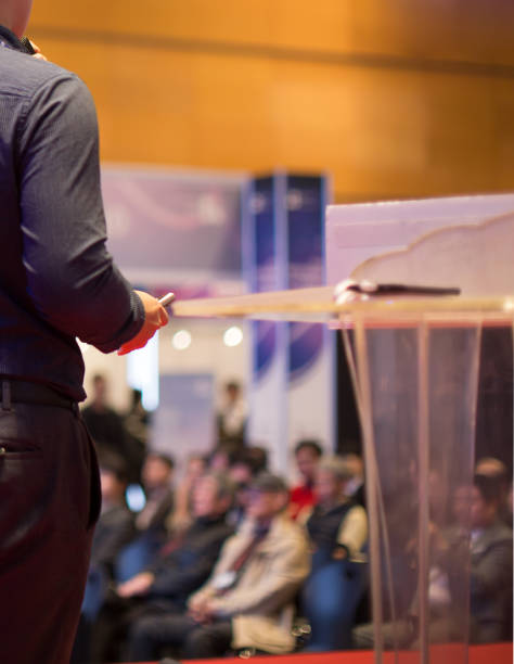 istock Focus on Remote Control and Hand of Presenter on Stage. Speaker Behind Podium in front of Audience at Conference Presentation. Attendees at Investor Event Hearing Investor Pitch at Seminar Meeting 1077646130