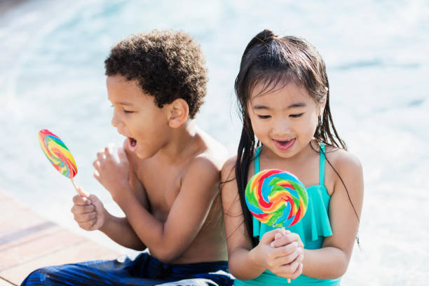 istock Two multi-ethnic children sitting by pool with lollipops 1077646140