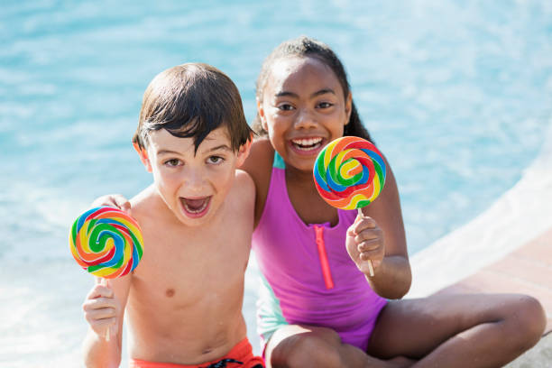 istock Two multi-ethnic children sitting by pool with lollipops 1077647340