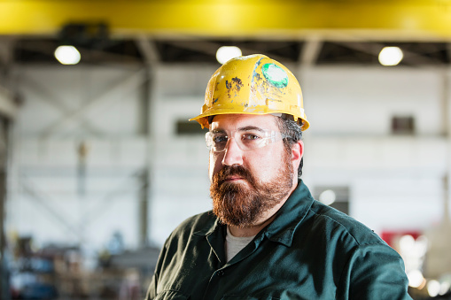 A mature man in his 40s wearing a hardhat and safety goggles, working in a manufacturing facility specializing in metal fabrication.  He is looking at the camera.