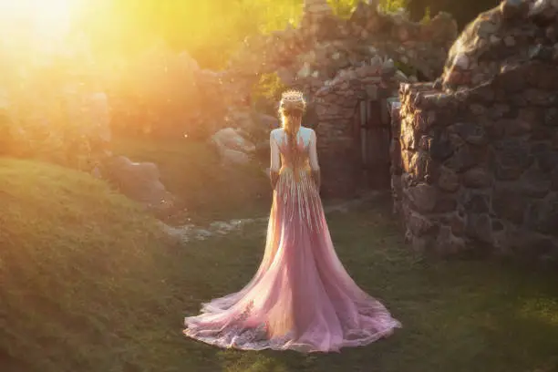Shooting without a face, from the back. Wonderful princess with blond hair and a crown. is wearing an amazing light pink dress with gold ornaments and a long train. Queen walks in the garden in the rays of sun.