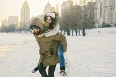 Theme New Year Christmas Mood Winter Snow Holidays Valentine Day. Young Caucasian couple lovers joy, laughter fooling water in city park. man holds woman on shoulders as backpack