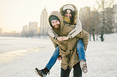 Theme New Year Christmas Mood Winter Snow Holidays Valentine Day. Young Caucasian couple lovers joy, laughter fooling water in city park. man holds woman on shoulders as backpack
