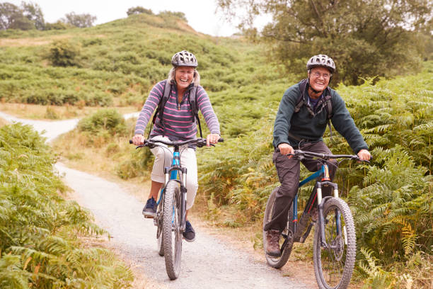 starsza para jeżdżąca na rowerach górskich w wiejskim pasie podczas wakacji kempingowych uśmiechnięta, widok z przodu - bicycle lane zdjęcia i obrazy z banku zdjęć