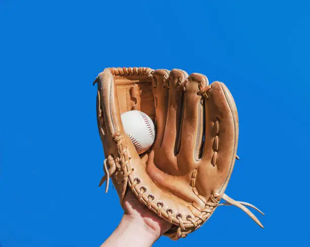 Photo of Hand in glove for a baseball game caught a leather white ball on a blue sky background. Sports contests. Victory. Achievement of success.