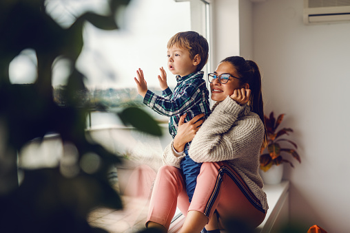 Mother sitting on the widow and holding her son. They looking through window.